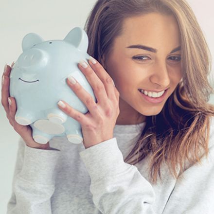 Eine Frau hält ein hellblaues Sparschwein mit beiden Händen neben ihrem Gesicht. Das Sparschwein hat ein lächelndes Gesicht und die Frau hat lange, braune Haare. Die Frau lächelt.